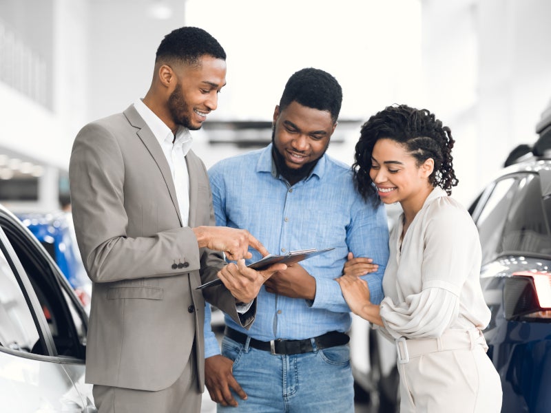 A couple inside a dealership, standing next to vehicles while talking to a sales rep, who is showing them something on his tablet